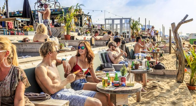Een gezellig druk terras met mensen in badkleding met hun voeten in het zand bij strandtent Indigo op Scheveningen in Den Haag Een gezellig druk terras met mensen in badkleding met hun voeten in het zand bij strandtent Indigo op Scheveningen in Den Haag